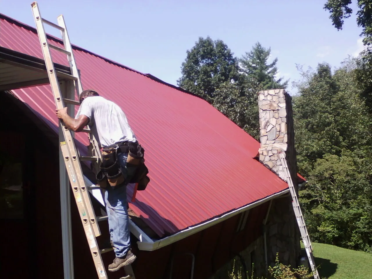 Roofer on a ladder beside a red metal roof and stone chimney — Soffit & Fascia Repair by Roofing Brothers in Perham
